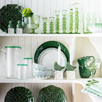 Puro mug in basil green, displayed among various green glassware and ceramics on a white shelf.