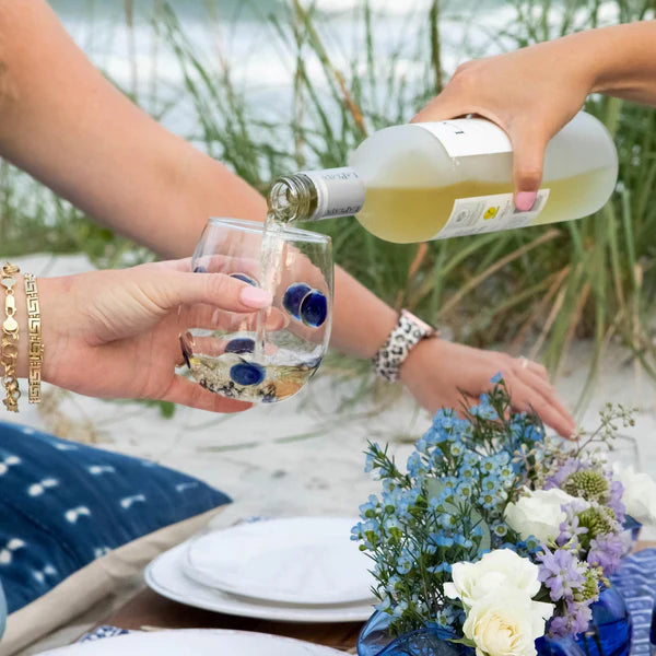 Drop blue stemless wine glass being filled with white wine, surrounded by a beach setting and floral arrangement.
