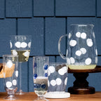 Drop blue tall tumbler with white polka dots, displayed alongside a matching pitcher and other glassware on a wooden surface.
