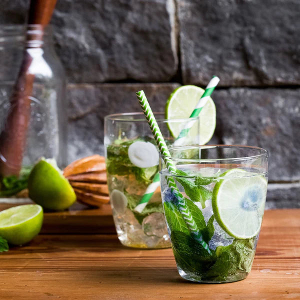 Drop blue tall tumbler with mint leaves, lime slices, and green striped straw on a wooden surface.