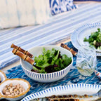 Sitio Stripe coupe bowl in Delft blue with a salad, placed on a patterned tablecloth with utensils and glassware.
